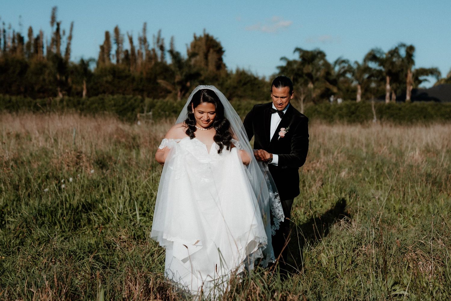 A wedding couple poses in a rustic field with tall grass and trees in the background.