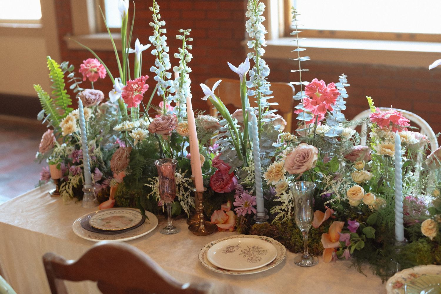 Table centerpiece with pink flowers, white candles, and greenery in rustic setting.