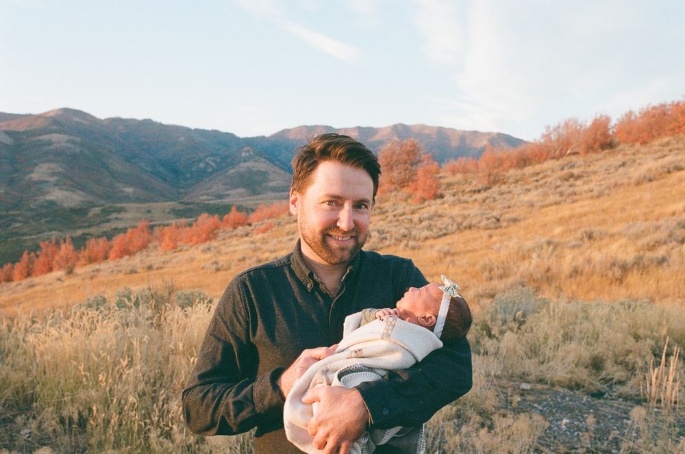 Man smiling outdoors holding newborn baby with autumn mountains and colorful foliage in background.