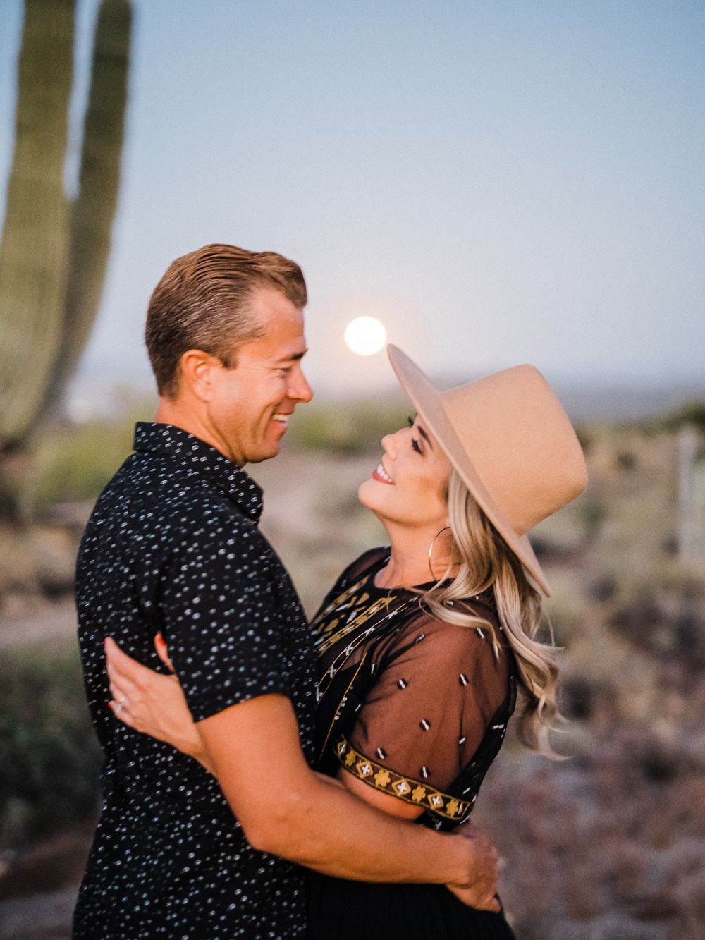 A couple embraces and smiles at each other in a desert landscape with saguaro cacti at dusk.