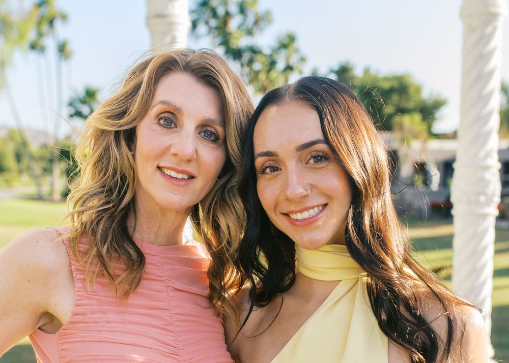Two smiling women in pastel dresses posing outdoors with palm trees in the background.