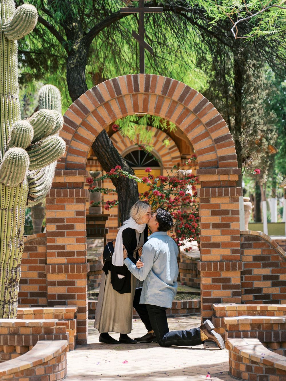 Couple sharing a romantic kiss under a brick arch surrounded by cacti and flowers in a garden.