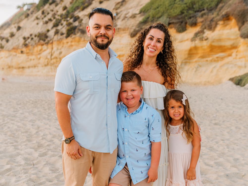 Happy family of four smiling together on sandy beach with scenic cliffs in background.