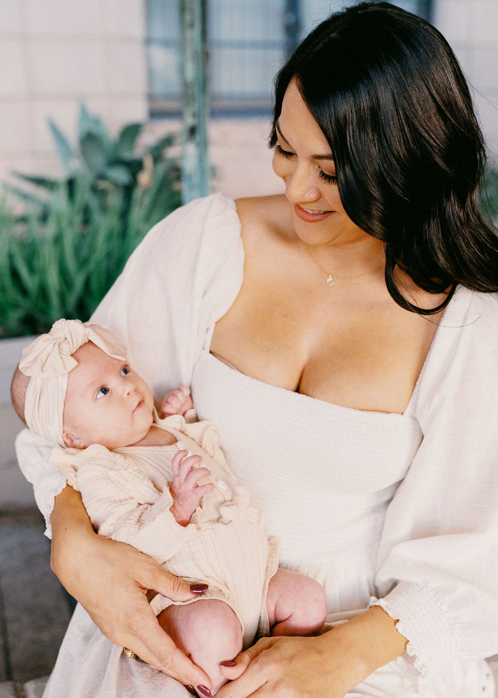 A dark-haired woman in a white dress tenderly holds her newborn baby dressed in a pink outfit.