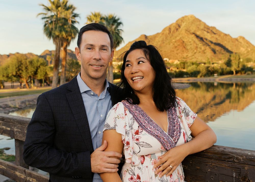 Couple smiling together outdoors near a lake with palm trees and a mountain at golden hour in Arizona.