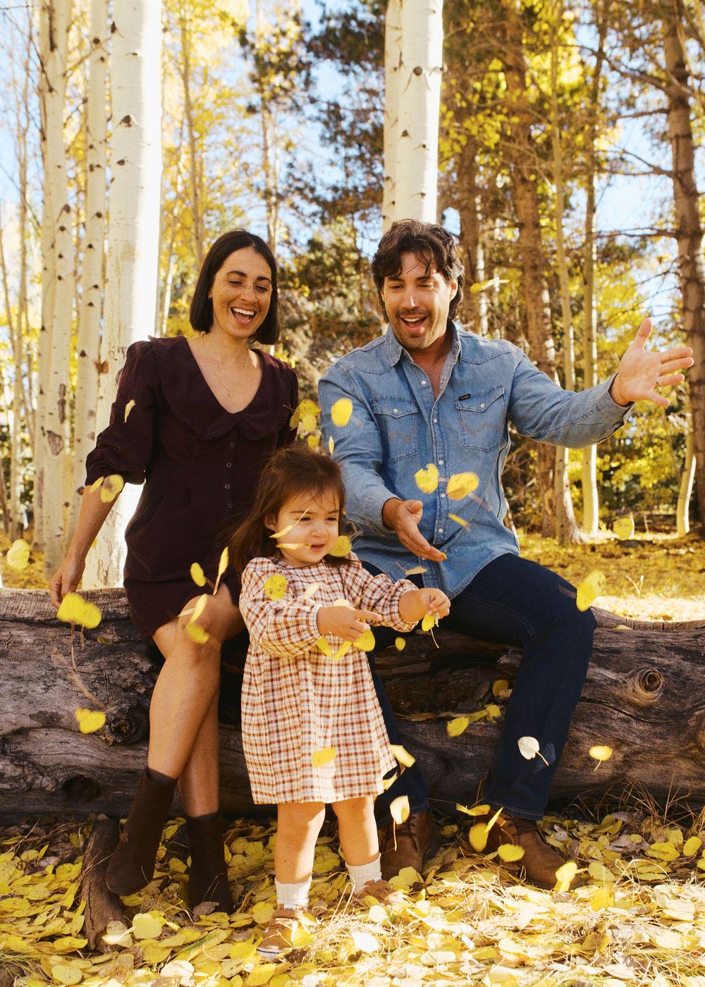 Family of three joyfully tossing yellow autumn leaves among aspen trees in a golden fall forest setting.