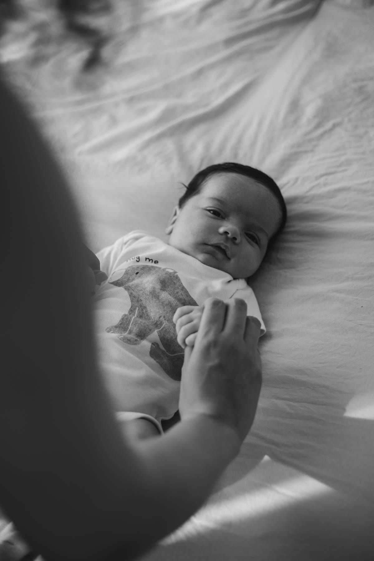 Black and white photo of a newborn baby lying on a bed while a parent gently holds the infant's hand.