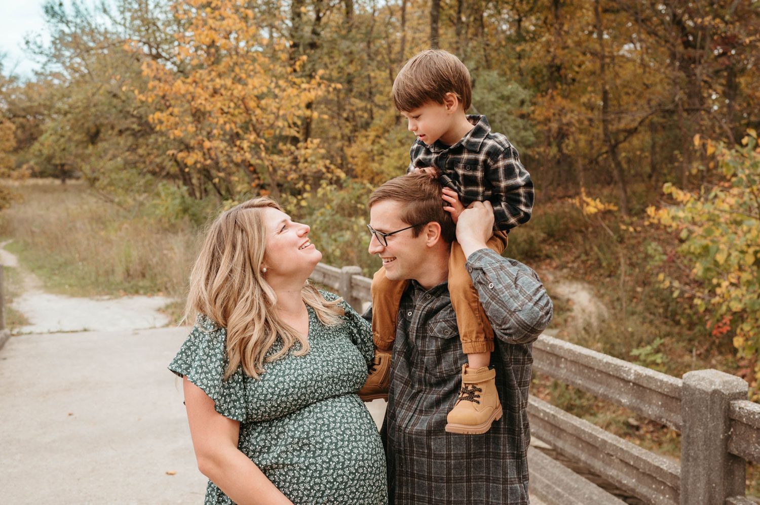 Pregnant woman and partner with child on shoulders on autumn path.