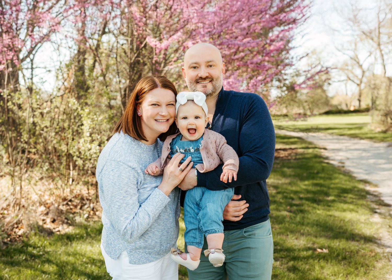 Smiling couple holding laughing baby girl in front of blooming pink redbud trees in spring park.