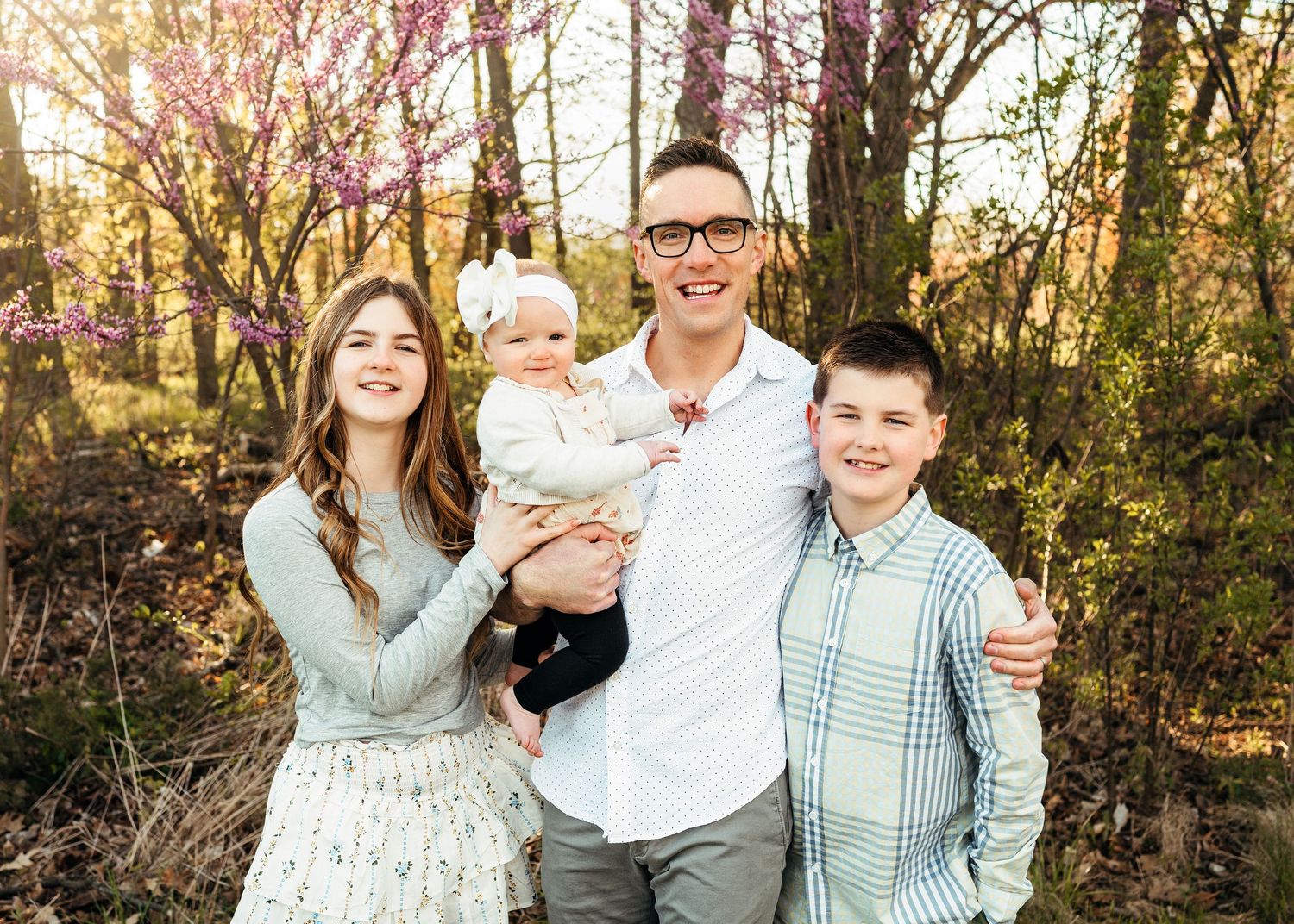 Family of four with parents holding baby girl and teen son smiling in spring woodland setting.