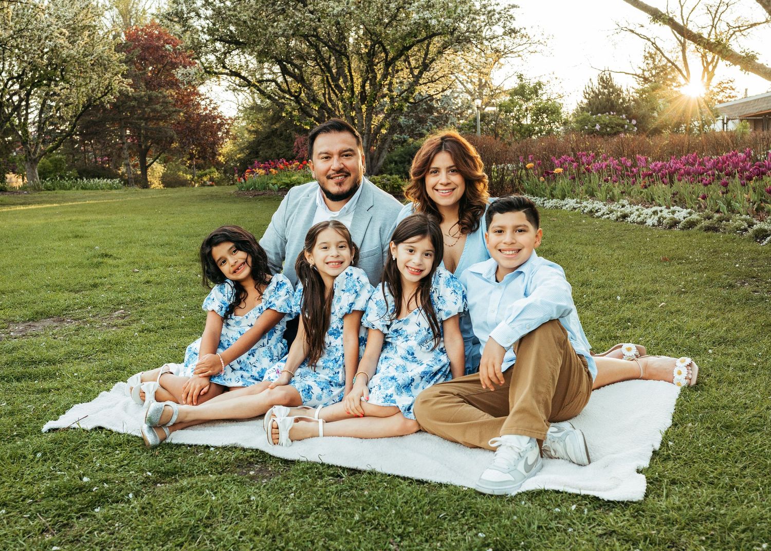Family of six in coordinating blue outfits posing on white blanket in sunny green park.