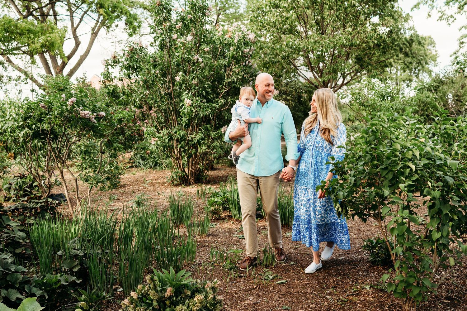 Family of three strolling through lush green garden path with father carrying baby in arms.