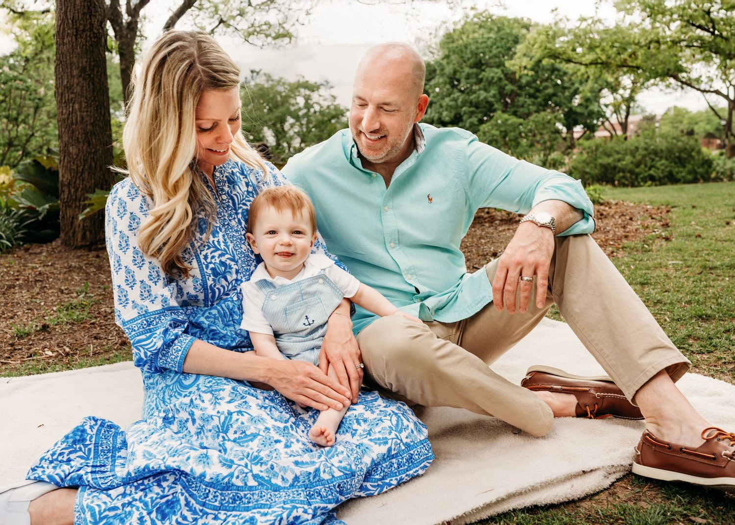 Young family with baby boy sitting on blanket in park, parents smiling down at child.