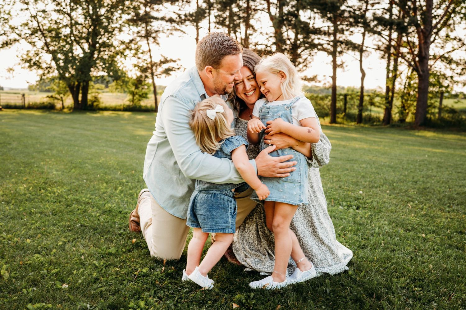 Happy family of four hugging and laughing together on green grass during golden hour outdoor session.