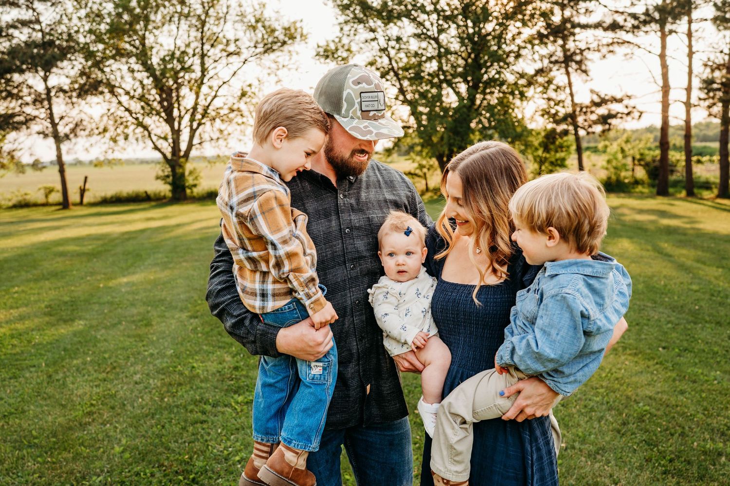 Family of five standing together on green lawn at sunset with parents holding young children.
