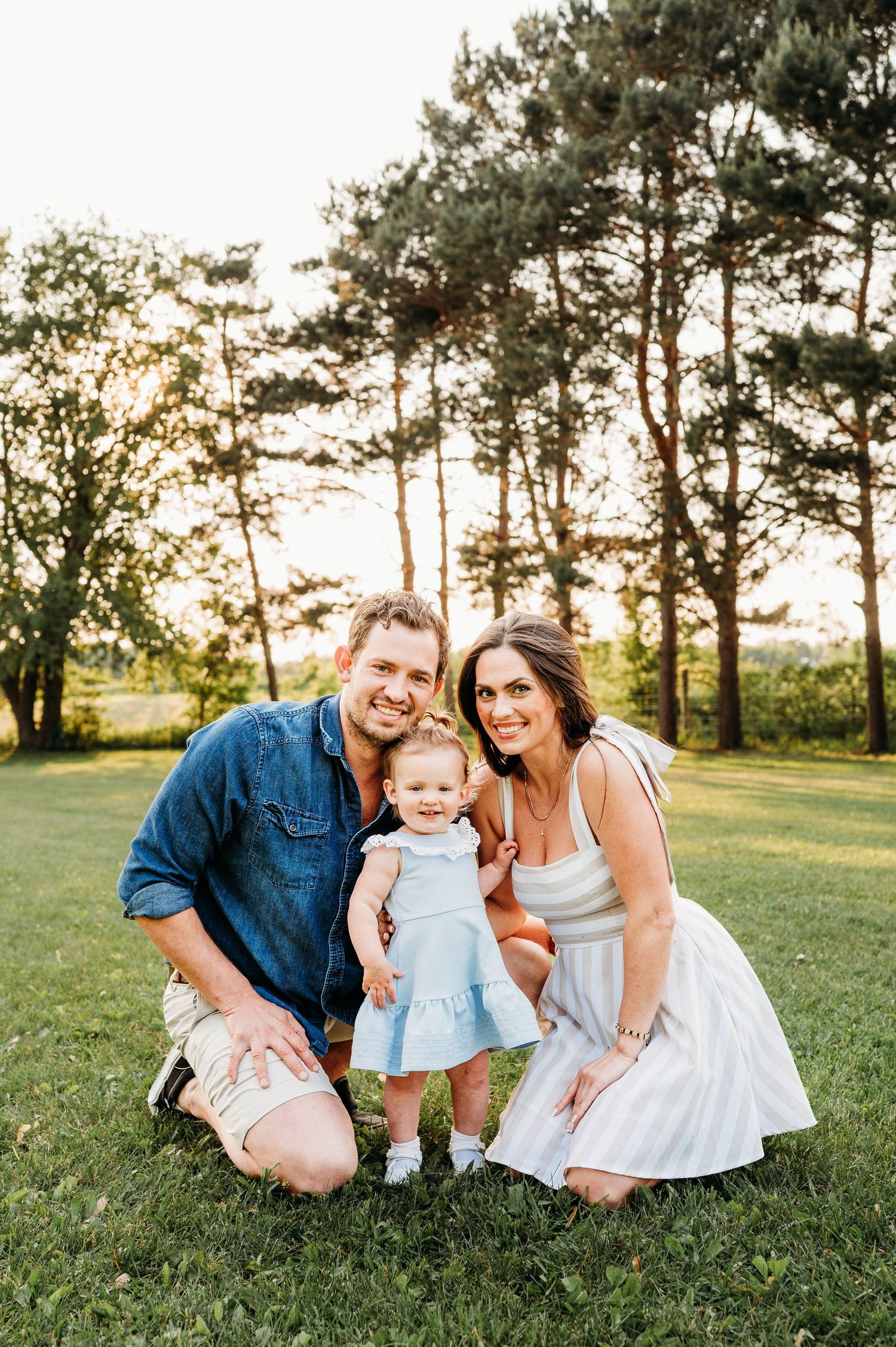 Smiling parents crouching with their baby girl in blue dress during golden hour outdoor family portrait.
