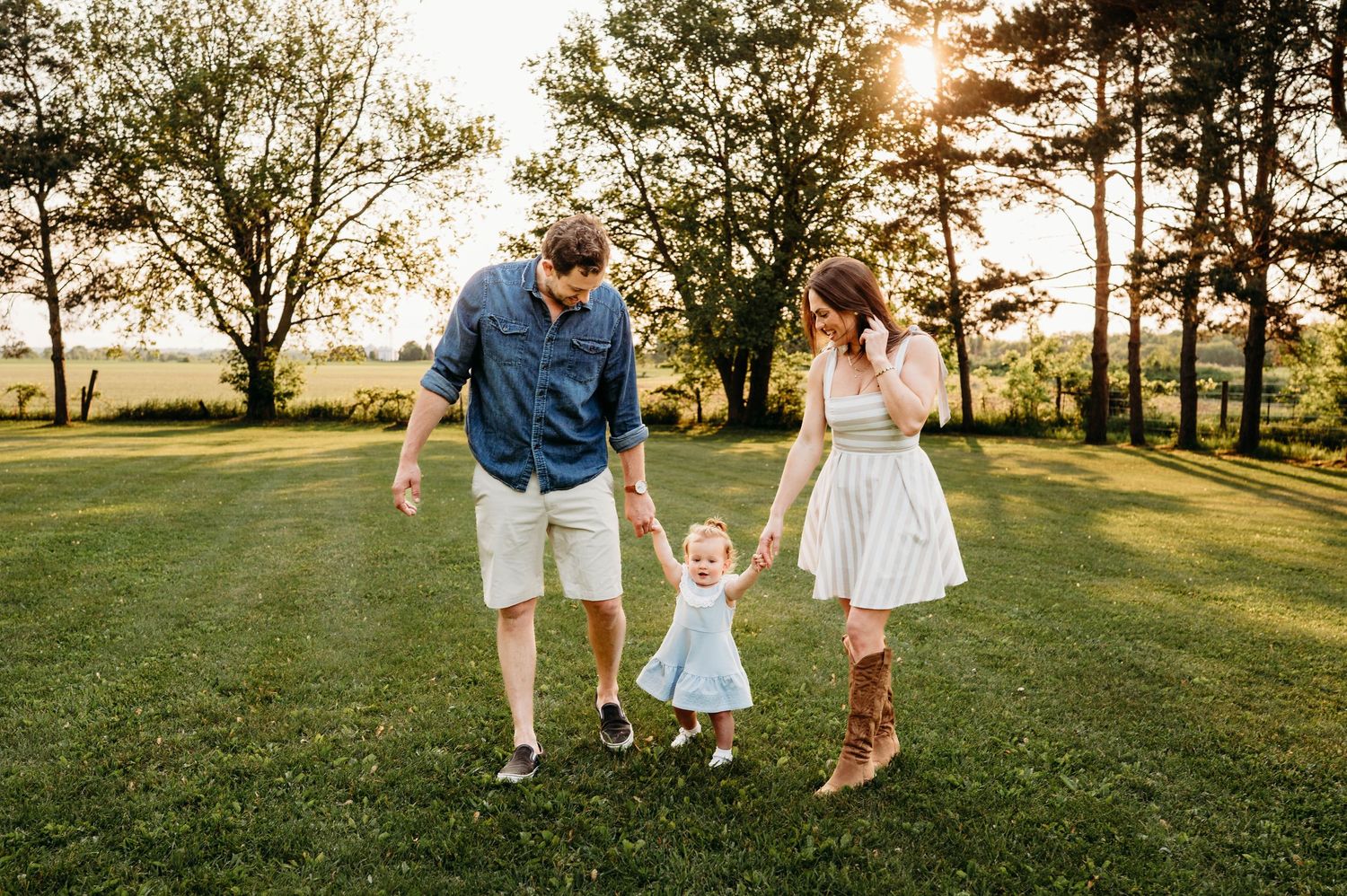 Young family walking together on green lawn holding toddler girl hands during sunny outdoor photo session.
