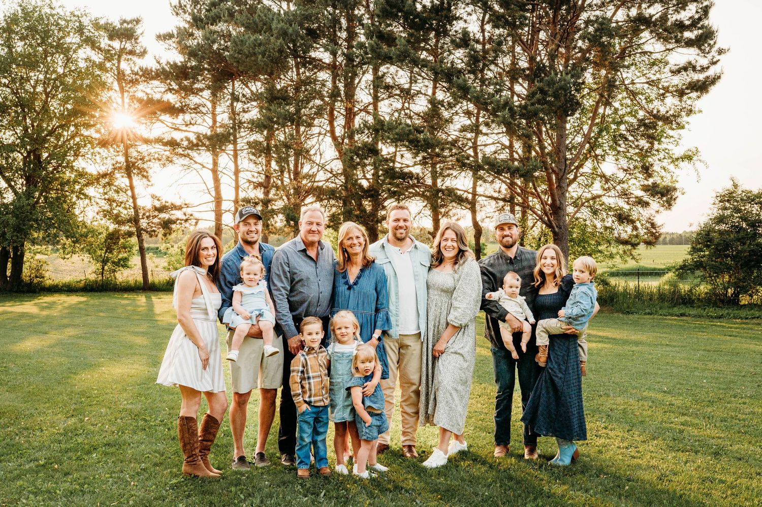 Large extended family smiling together on green lawn at golden hour with tall pine trees in background.