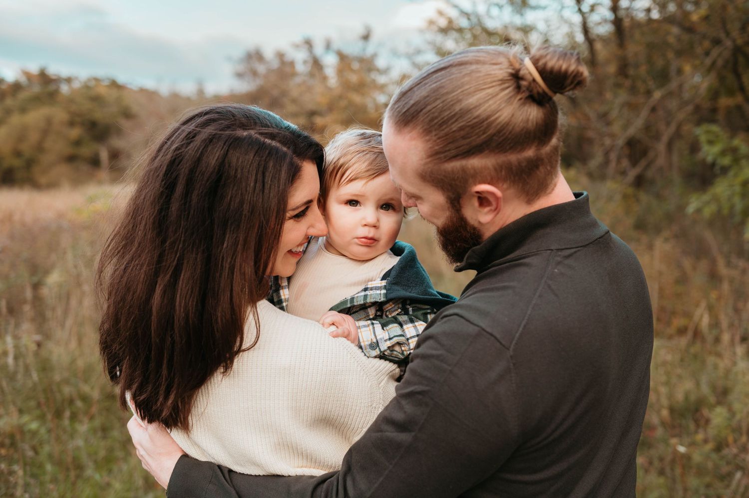 Young parents holding toddler in plaid jacket in autumn meadow.