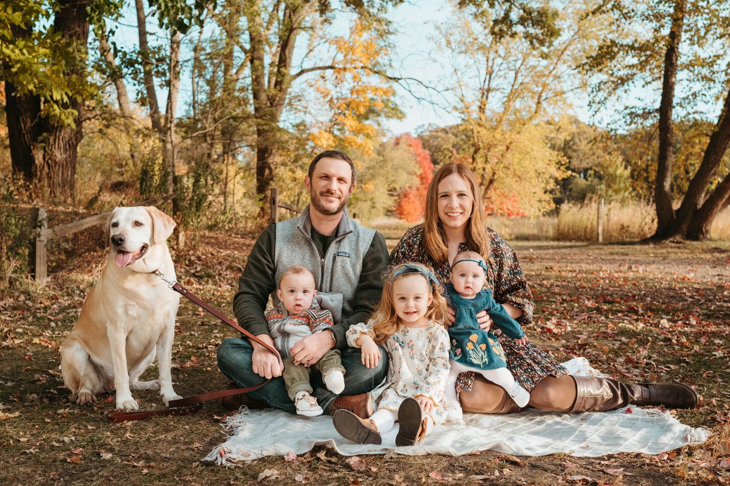 Family with three young children and yellow lab on blanket in fall foliage.