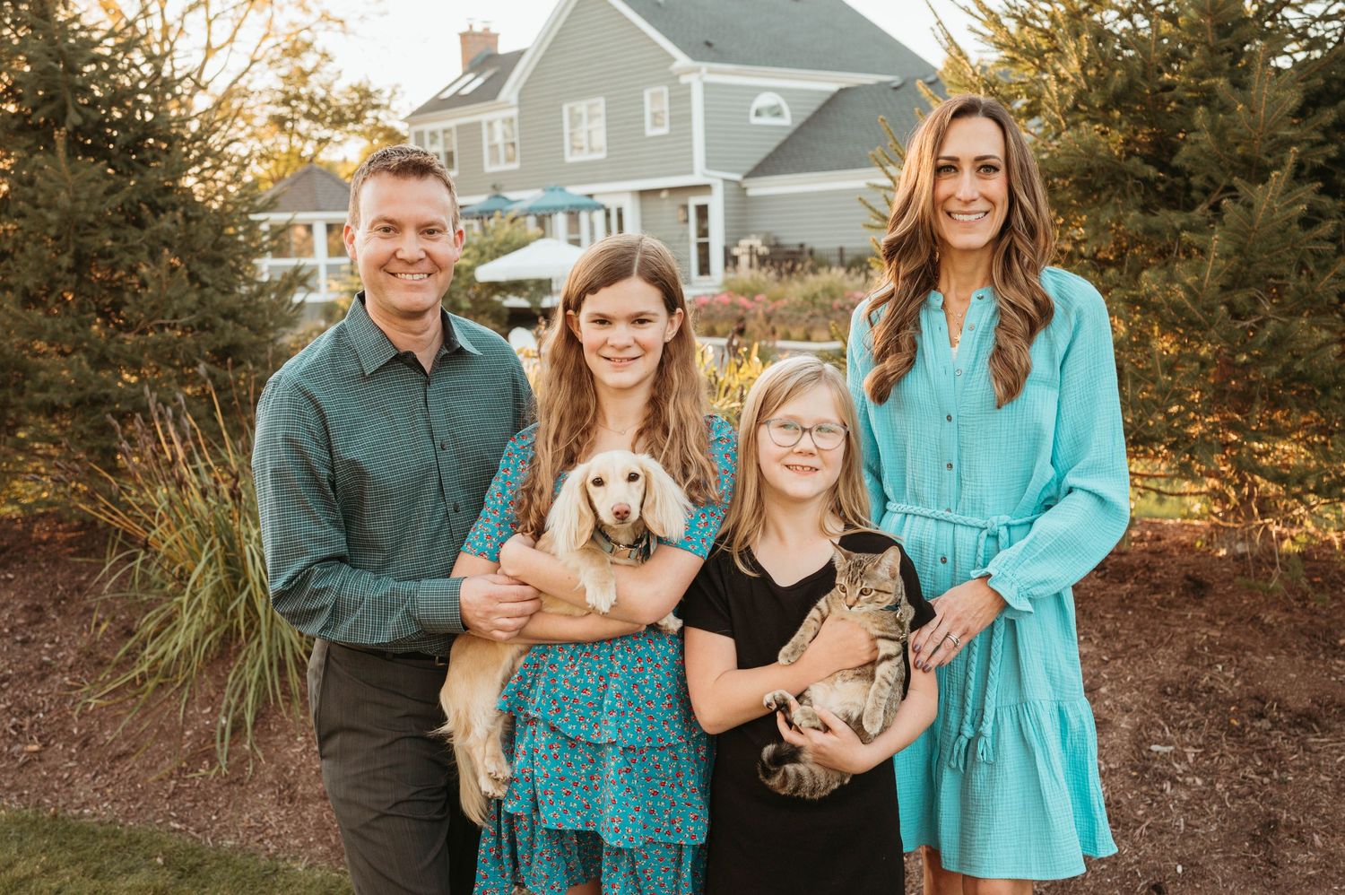 Family of four with dog and cat posing outside suburban home in teal outfits.