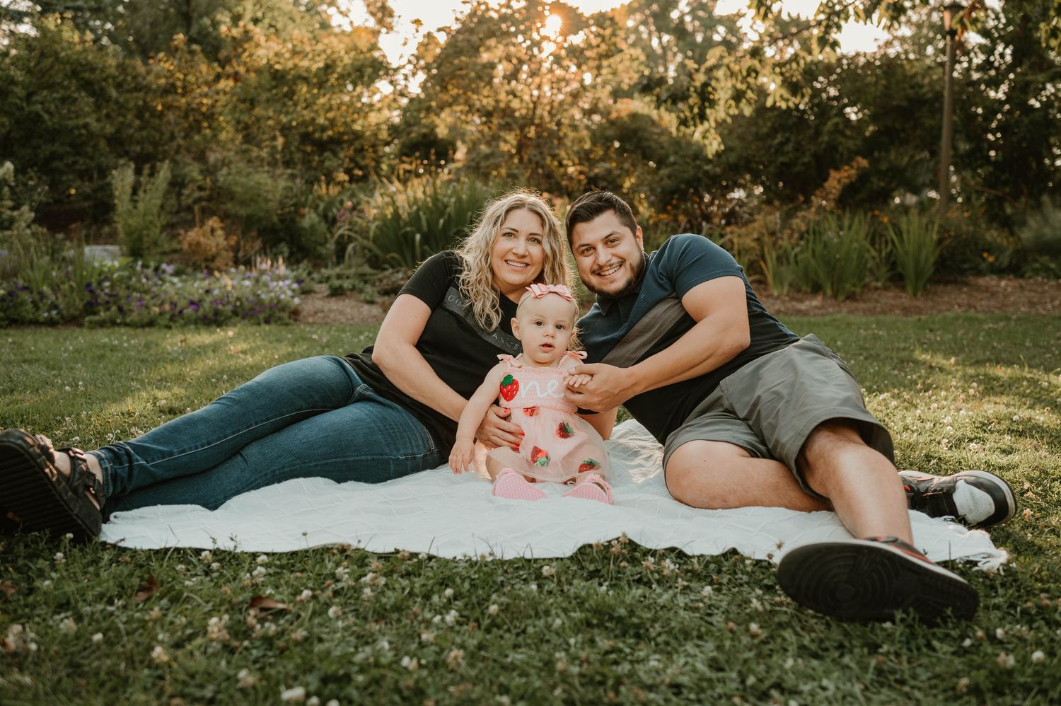 Happy family with blonde mom, dark-haired dad, and baby girl sit on white blanket in sunny park.