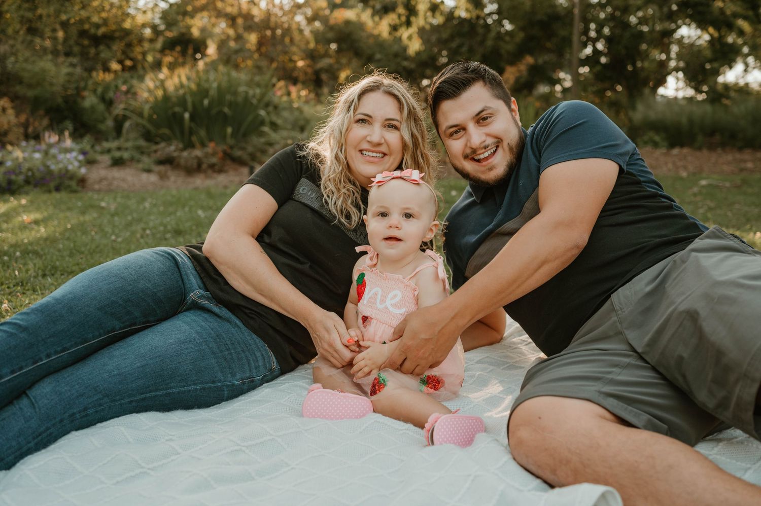 Smiling parents in casual black and navy outfits sit on blanket with their baby girl in pink dress.