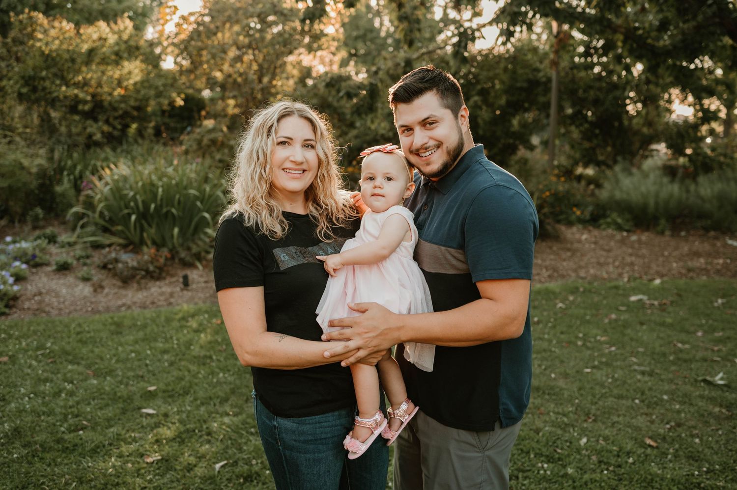 Smiling couple stands in garden holding their baby girl in a white dress during golden hour.