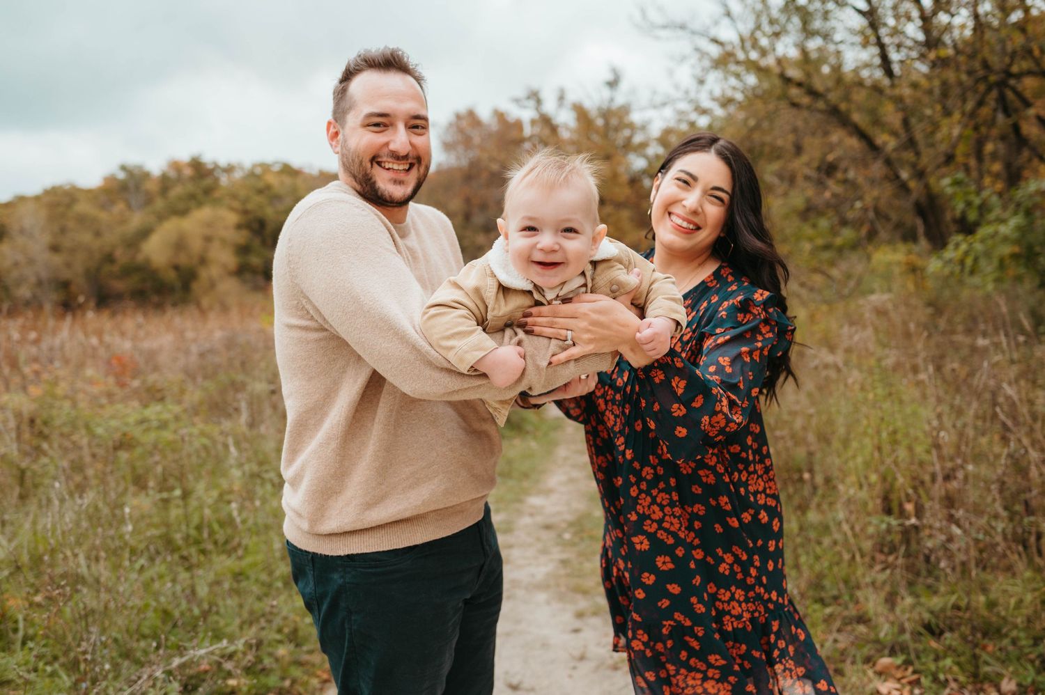 Parents holding laughing baby on scenic autumn nature trail path.