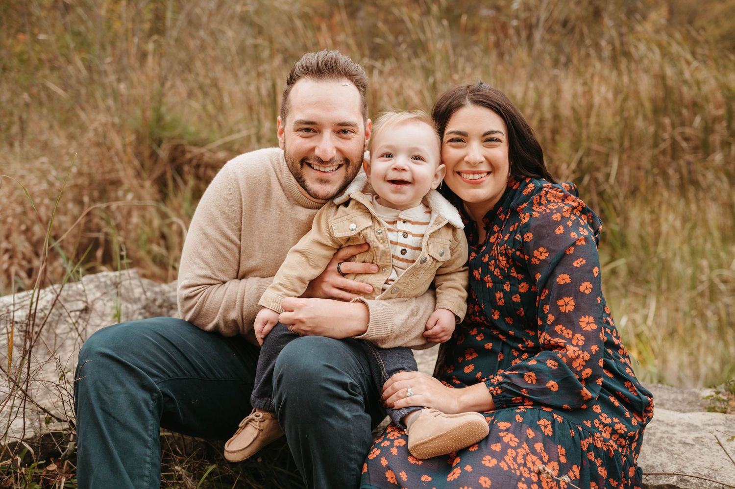 Smiling couple with happy baby sitting on rocks in autumn field.