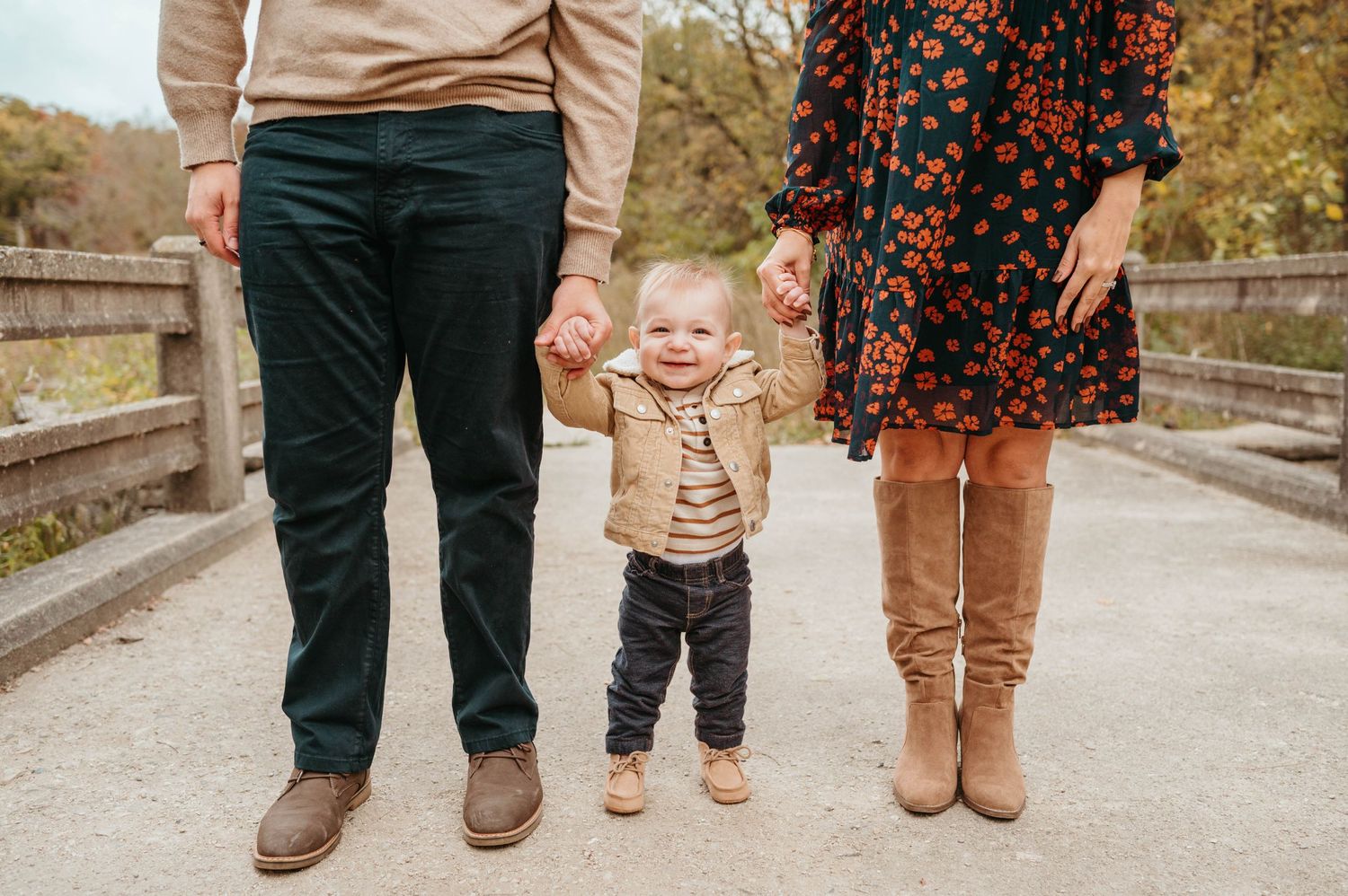 Toddler holding parents hands while standing on bridge in fall park.