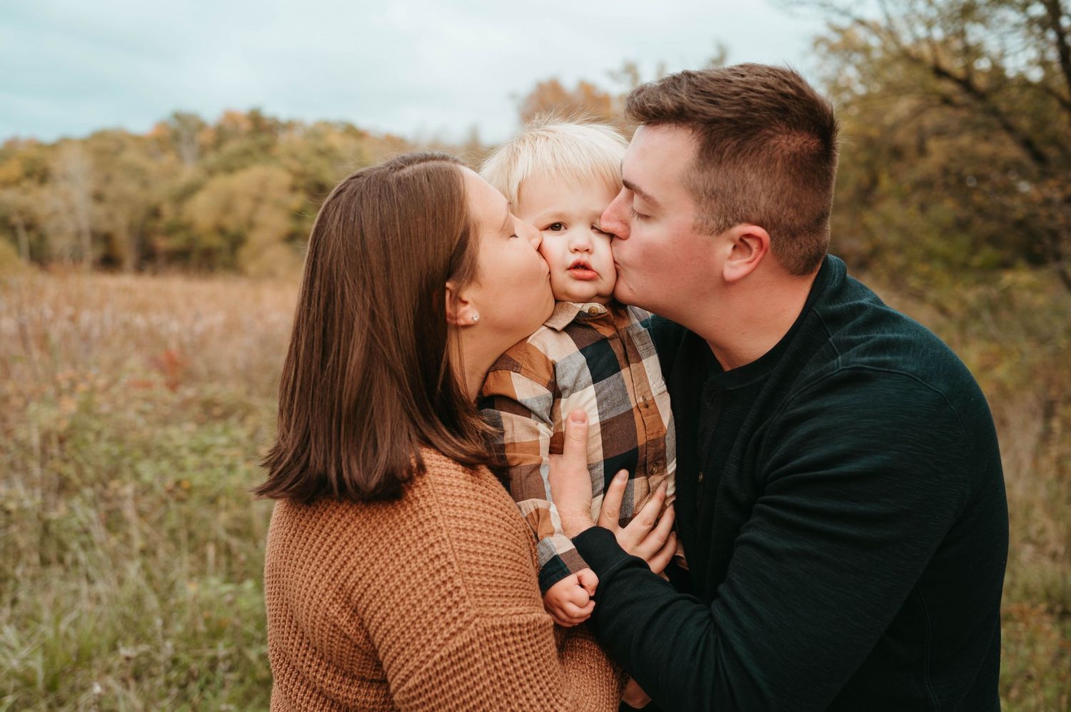 Parents kissing toddler on cheeks in autumn field portrait.