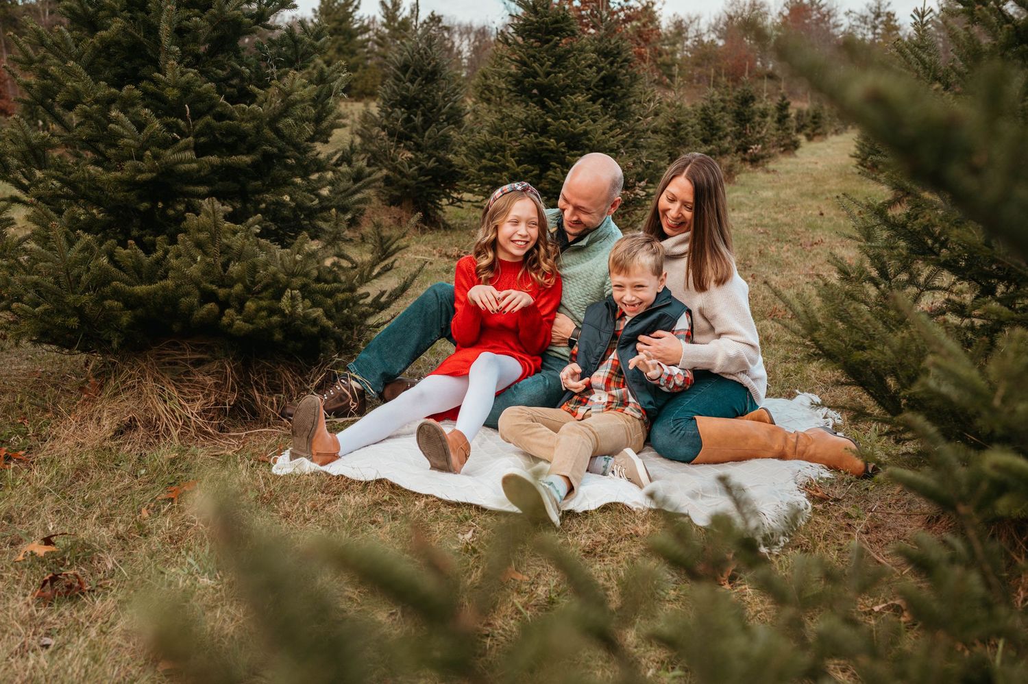 Happy family sitting on blanket among Christmas trees at outdoor tree farm.