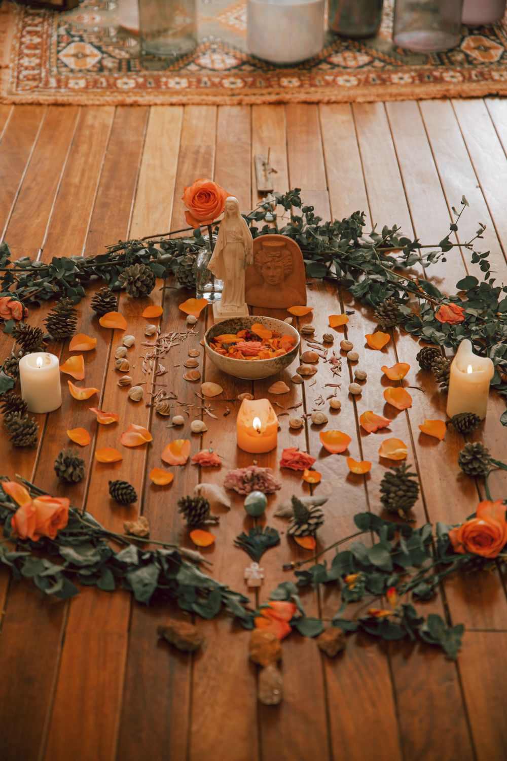 Romantic setup with orange roses, candles and greenery arranged on a wooden floor in a circular pattern.