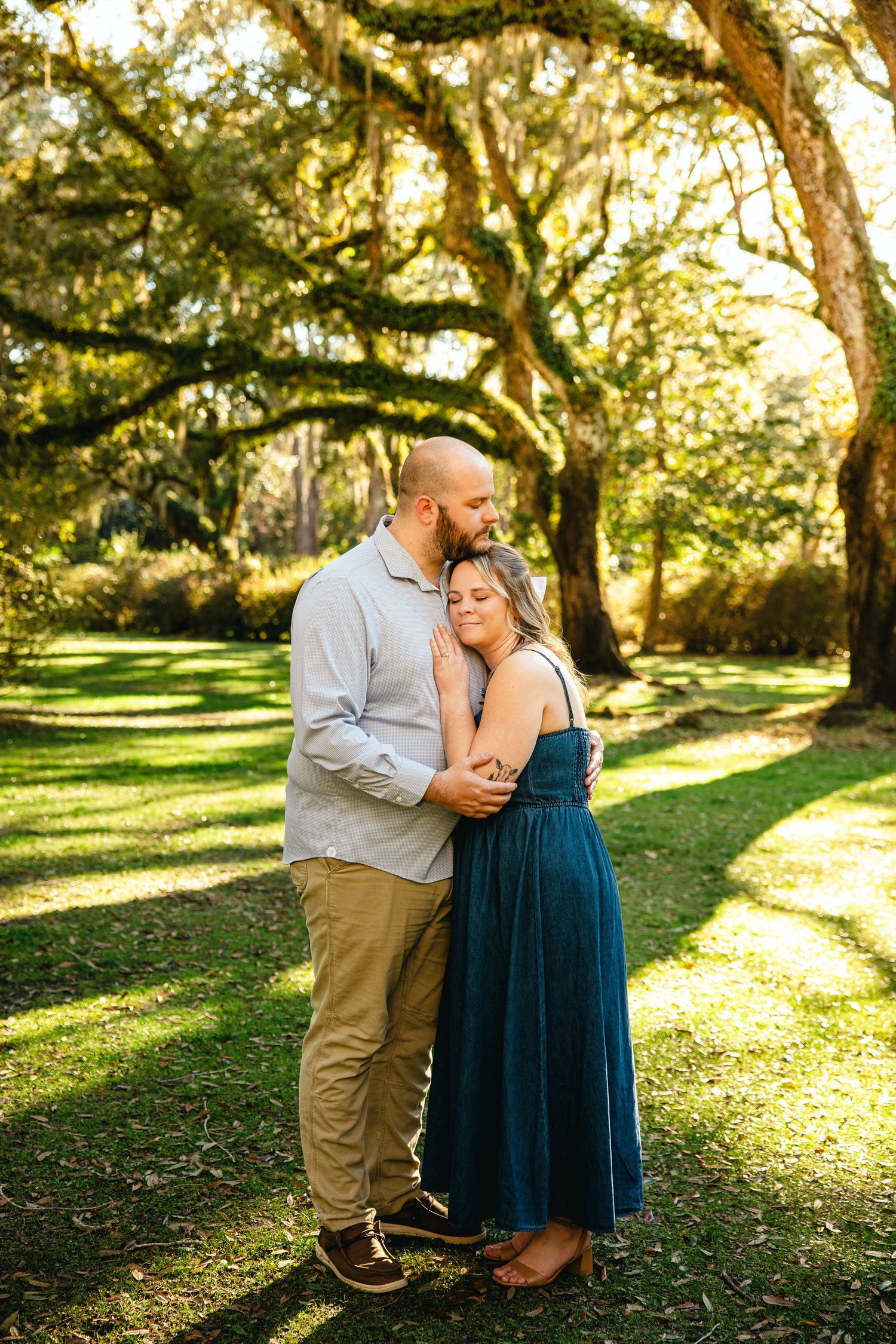 Couple shares intimate moment in sunlit park with moss-covered trees.