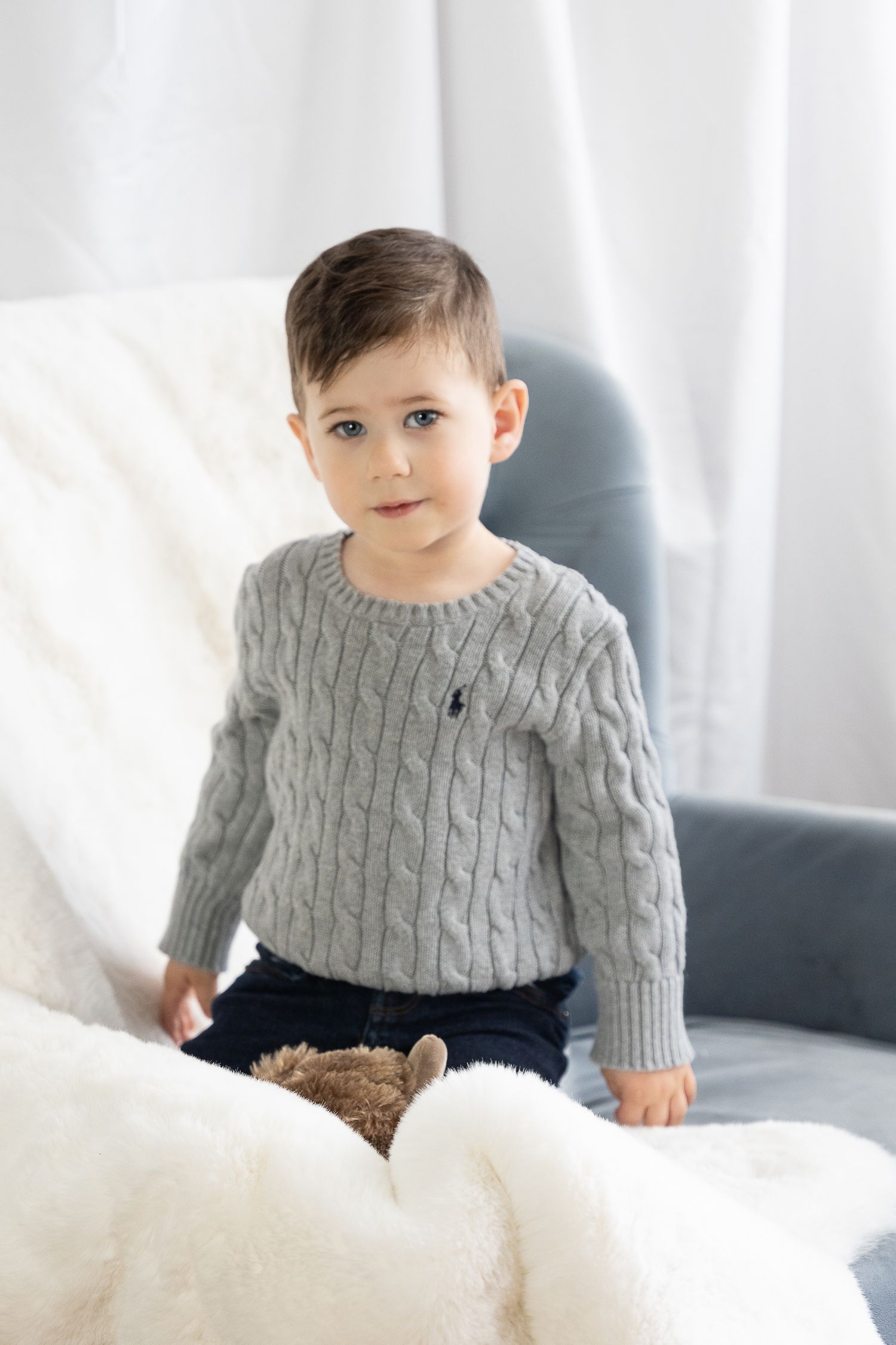 Young child in gray cable knit sweater sitting on white furniture in bright room.