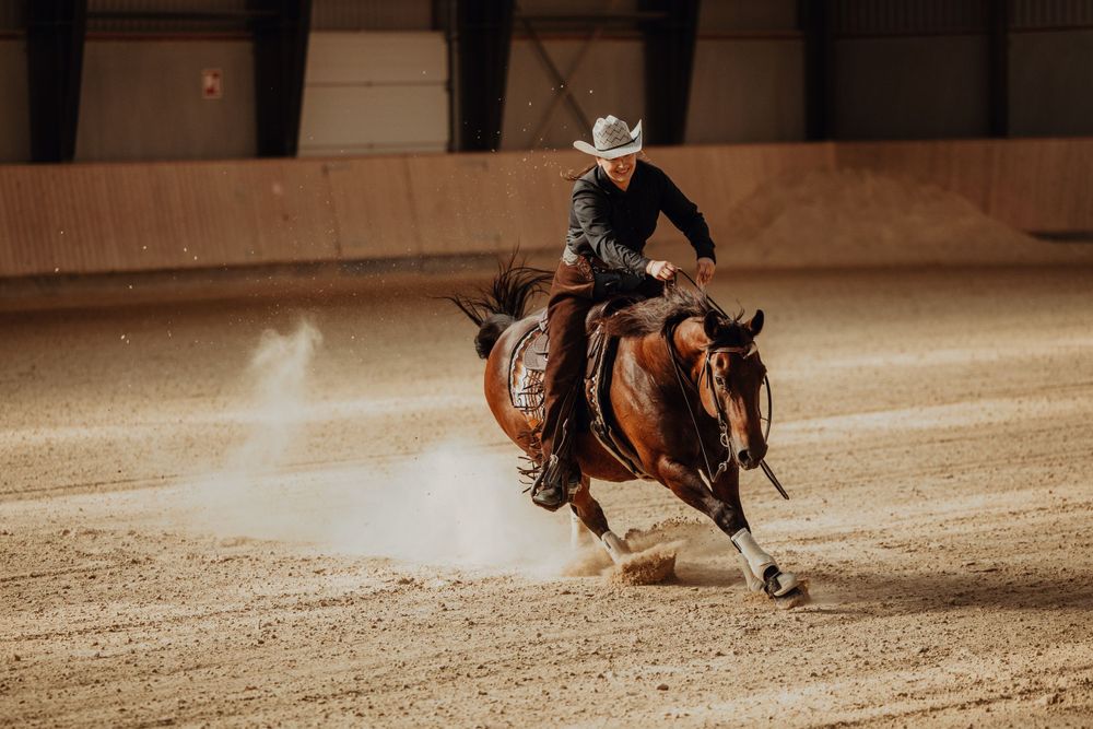 A horse and rider perform a skilled reining maneuver in a dusty indoor arena during competition.