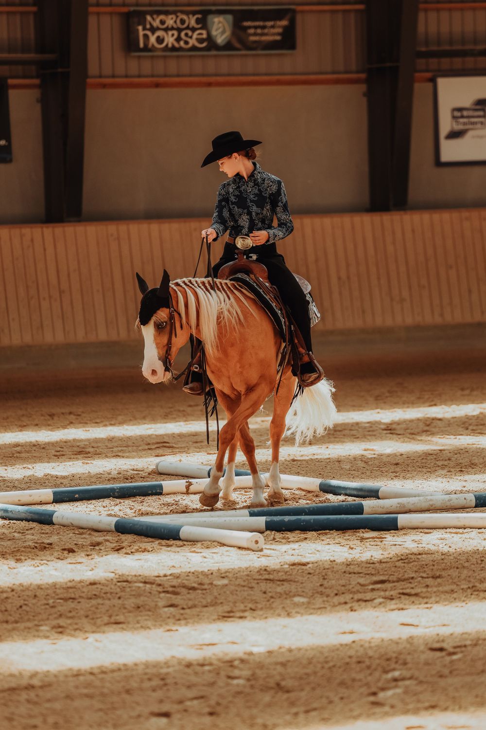 Western rider in black attire guides paint horse through pole obstacles during indoor arena training session.