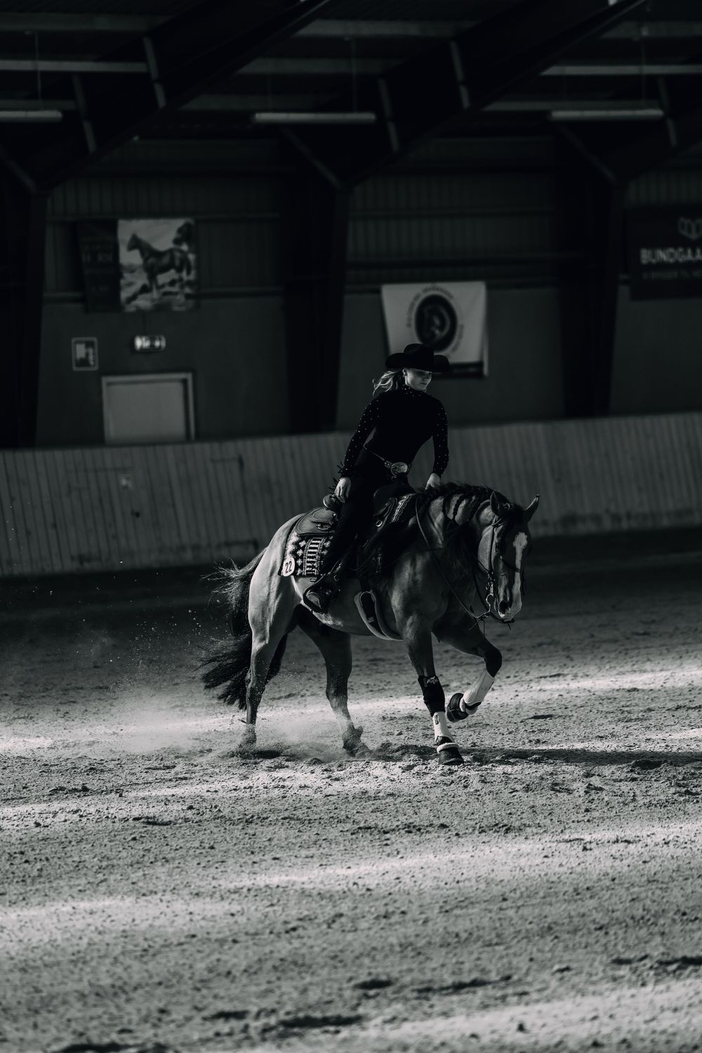 A rider in black performs dressage on a horse in an indoor arena with dramatic black and white lighting.