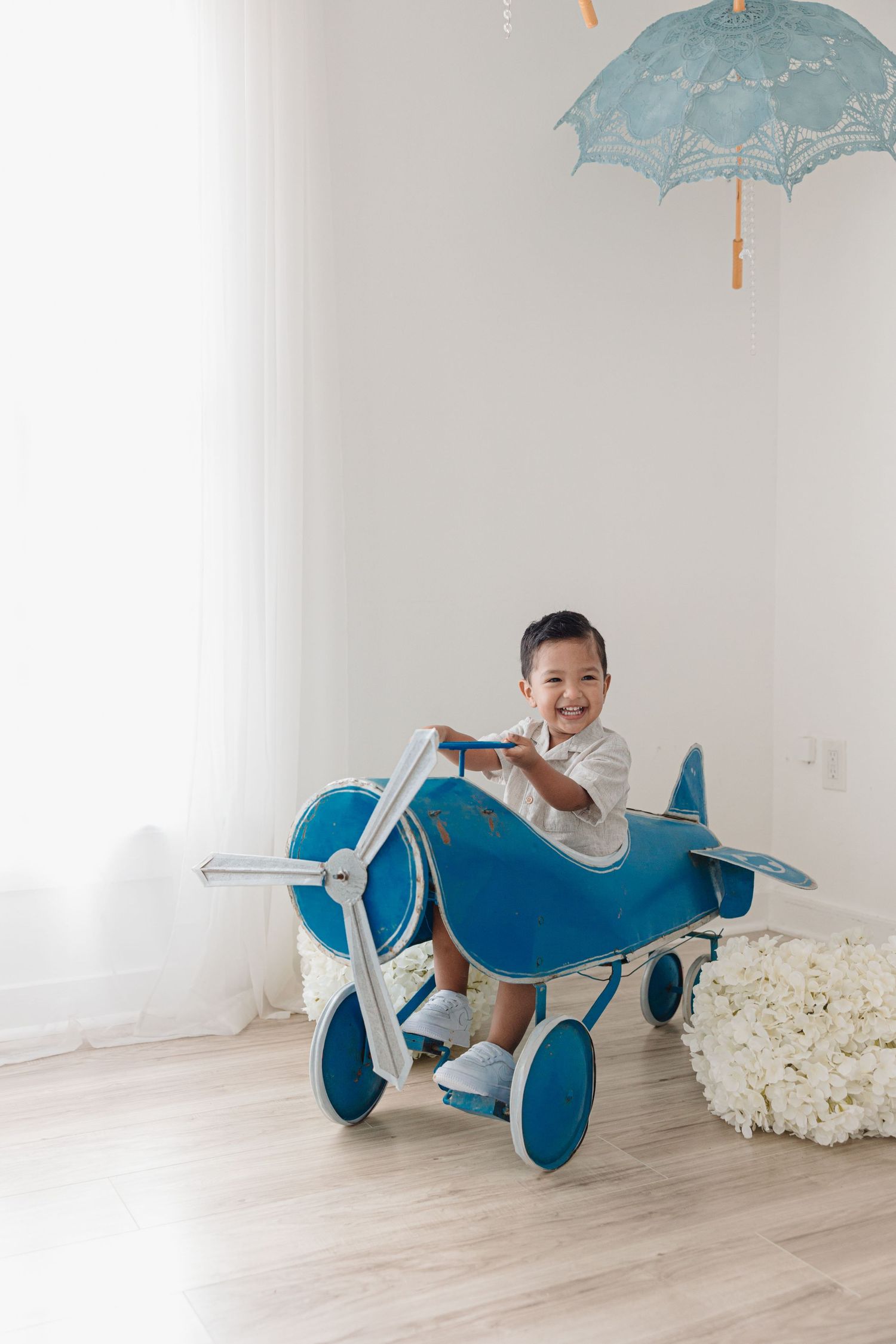 Child riding blue toy airplane under decorative umbrella in bright white room.