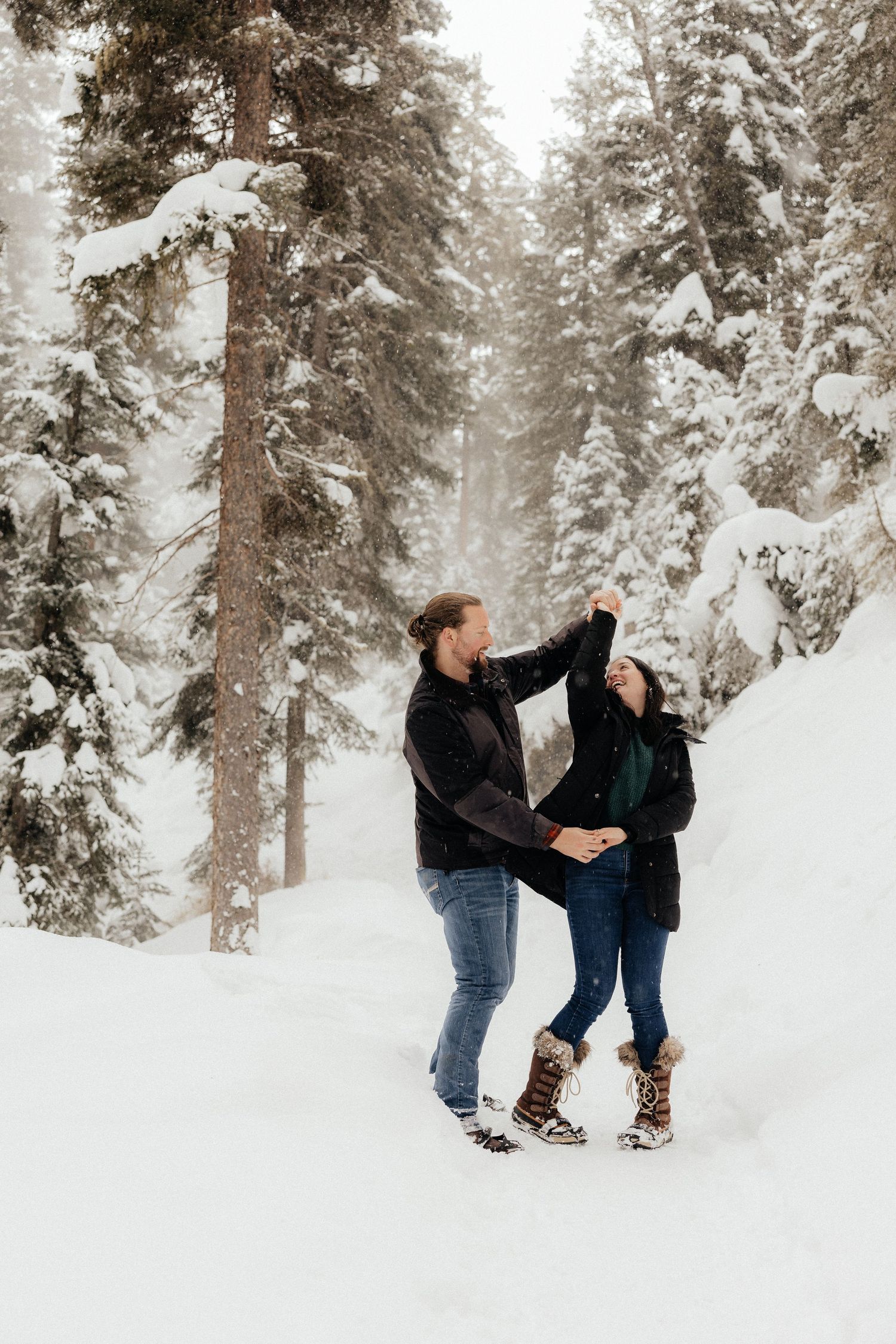 A couple enjoys a romantic winter moment together in a snow-covered forest with tall evergreen trees.