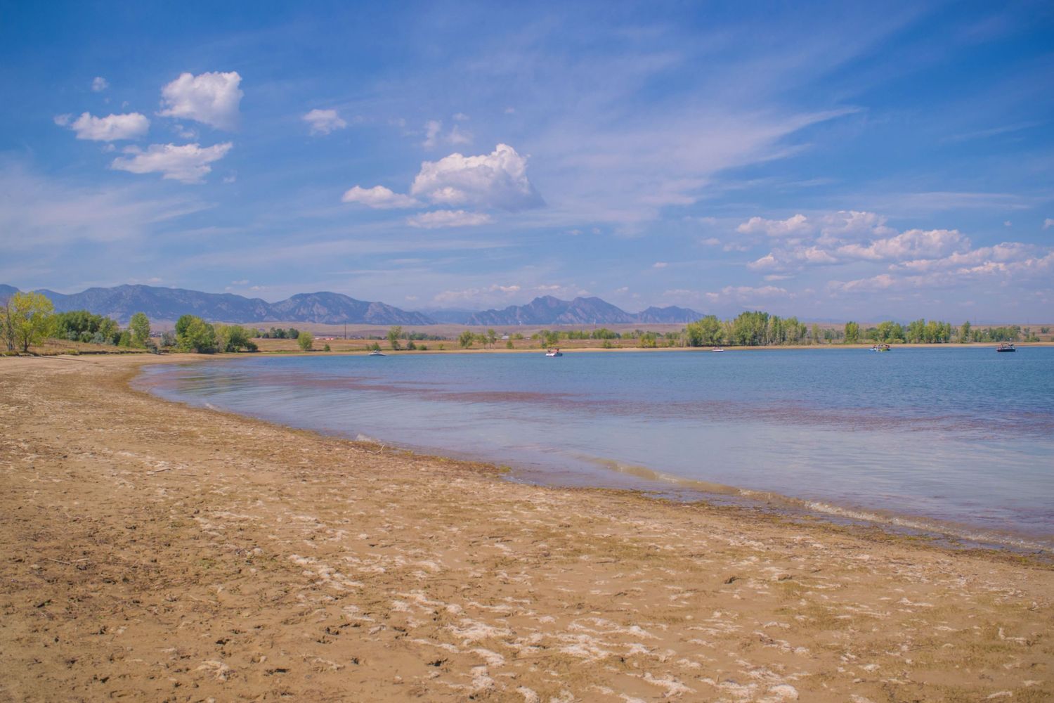 A scenic lake shore with sandy beach and calm blue waters against distant mountains on a sunny day.