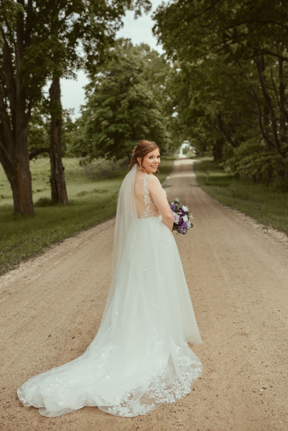 A bride in a lace detailed gown with train poses on a countryside path.