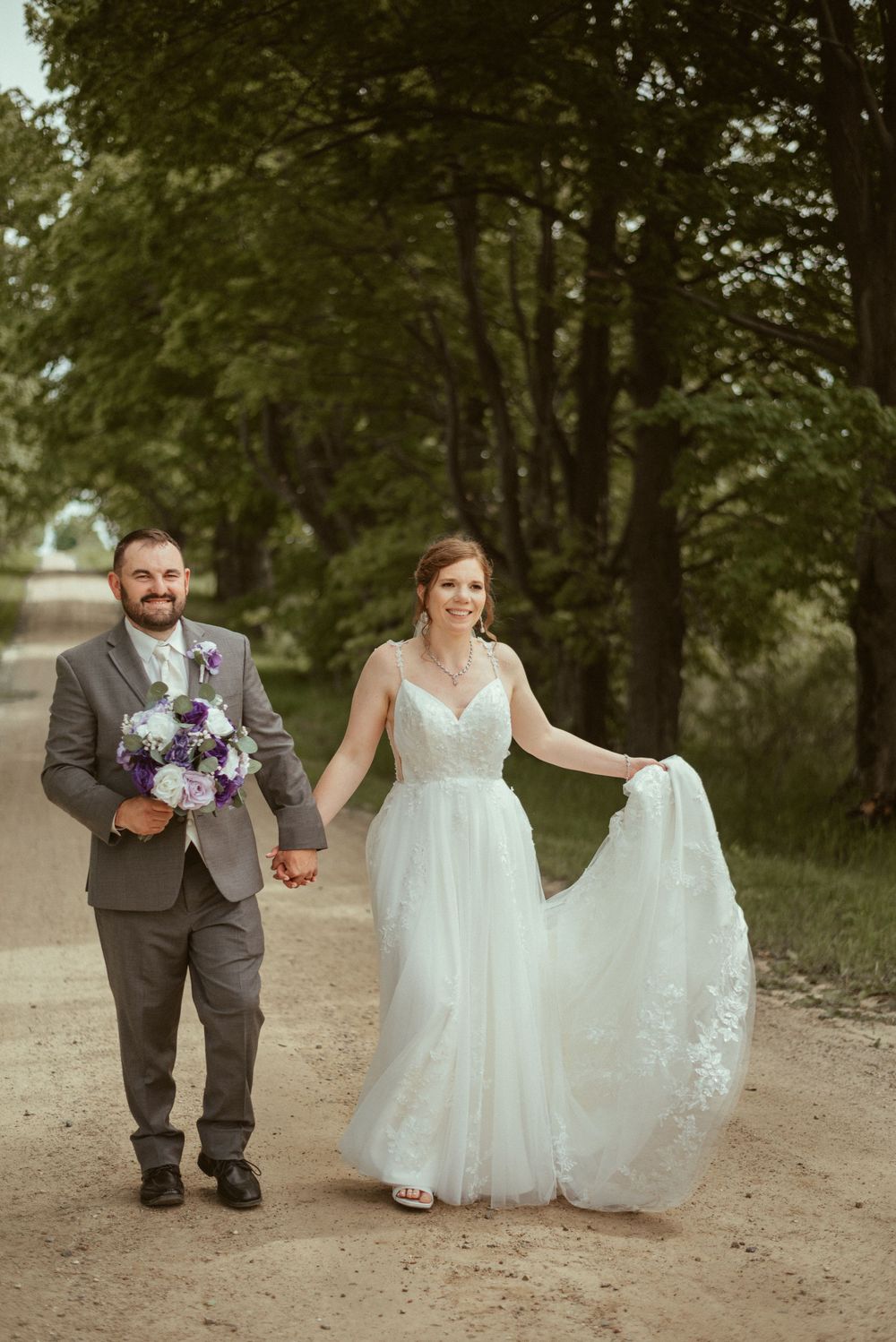 Newlywed couple walking down a path surrounded by trees as bride holds her flowing white dress.