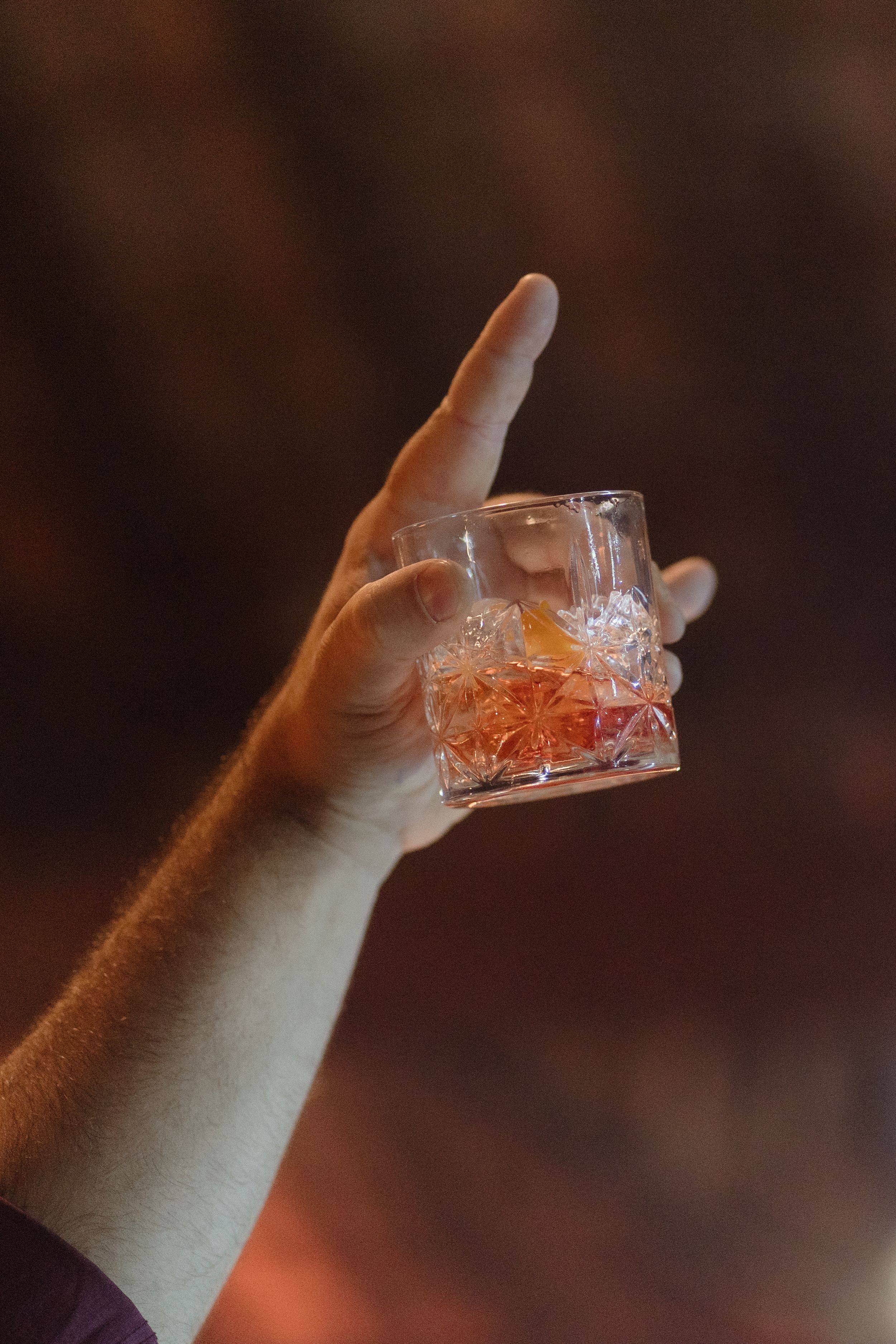 A hand holds up a crystal glass with amber-colored liquid against a dark background.