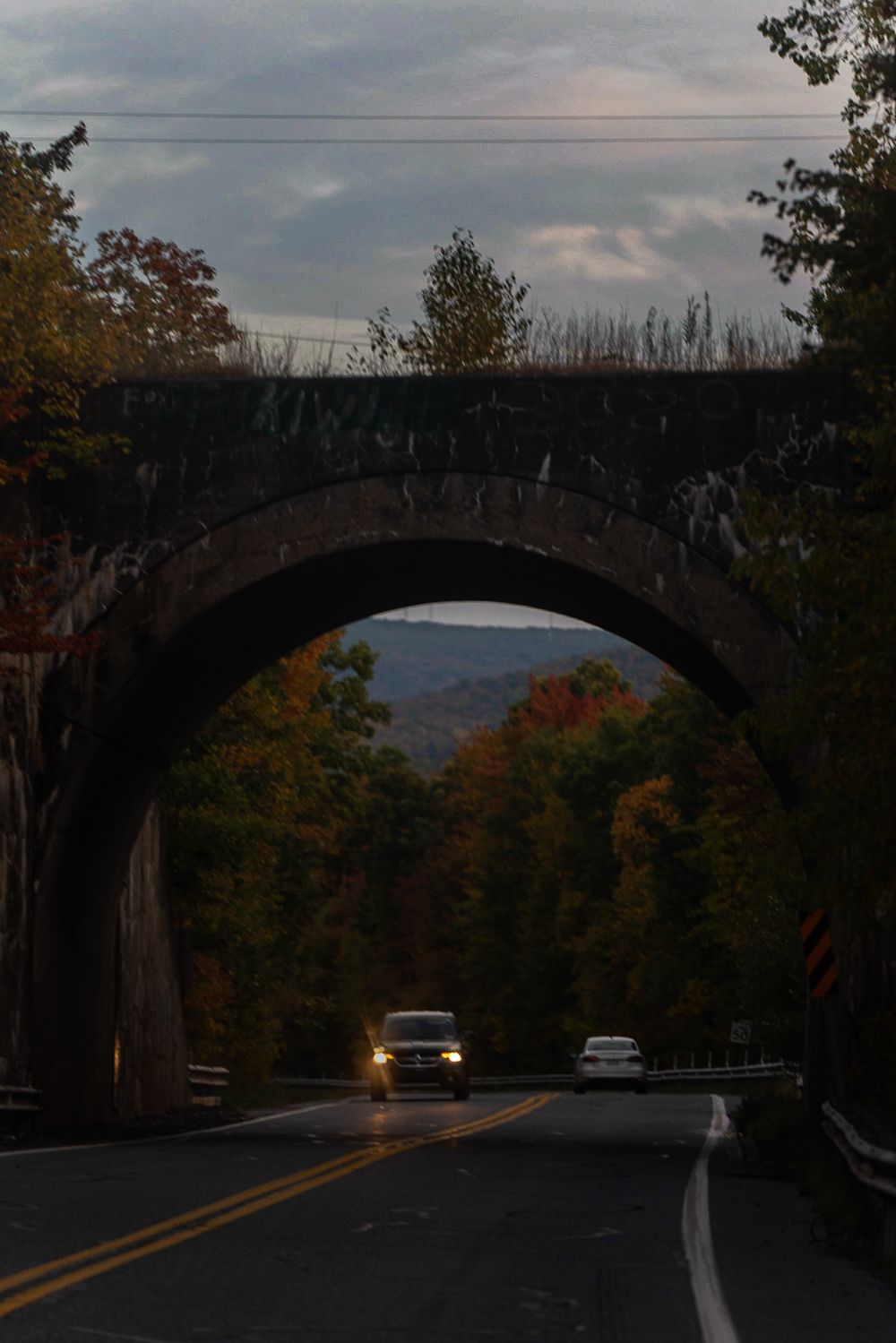 Stone arch bridge over a road with cars passing through during sunset and fall foliage.