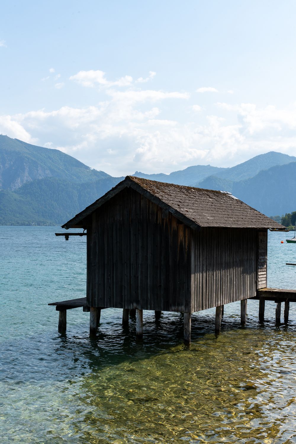 Wooden boathouse on stilts over pristine alpine lake with mountains in background.