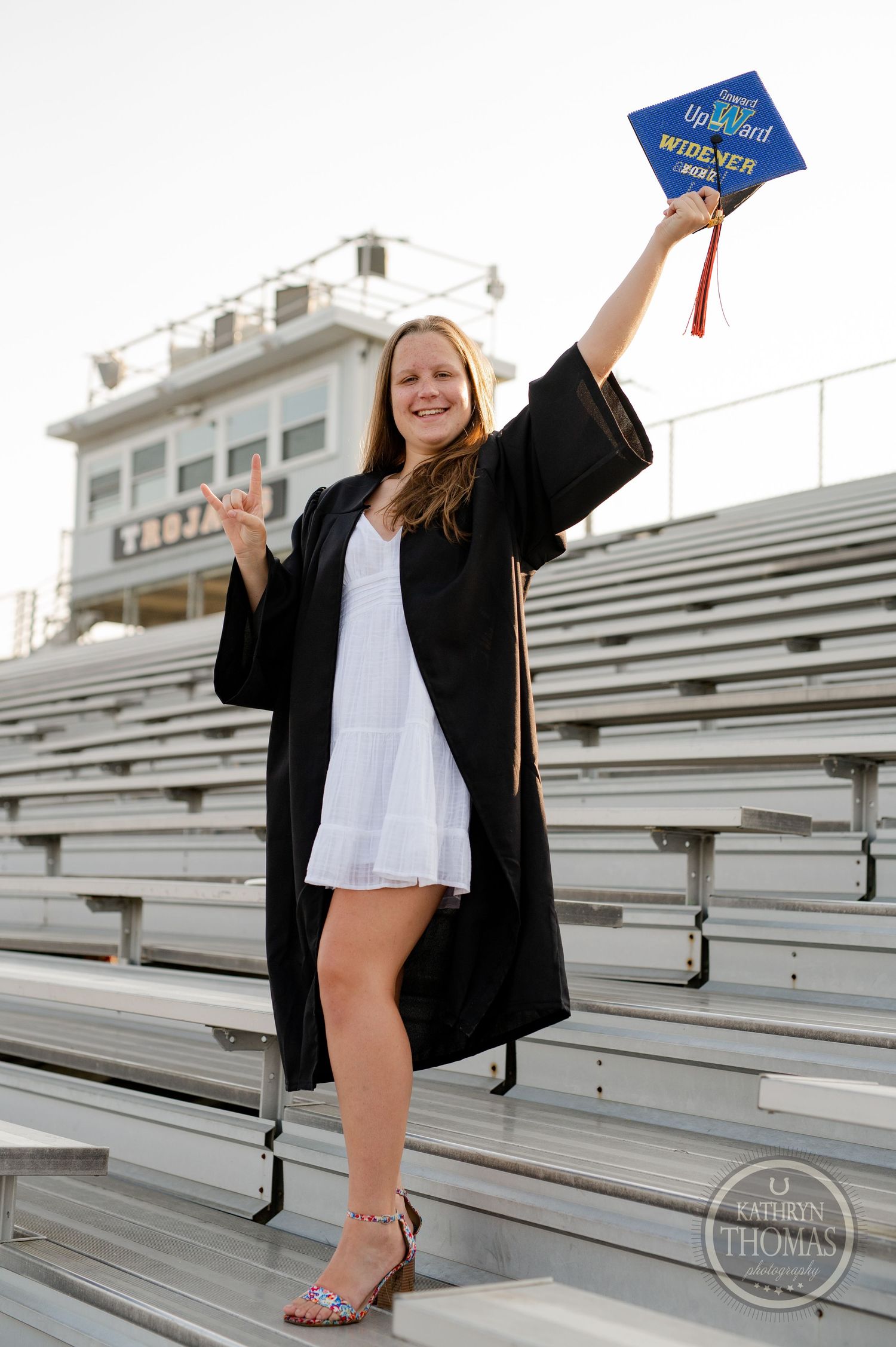 Graduate celebrates on bleachers holding decorated cap against stadium backdrop.