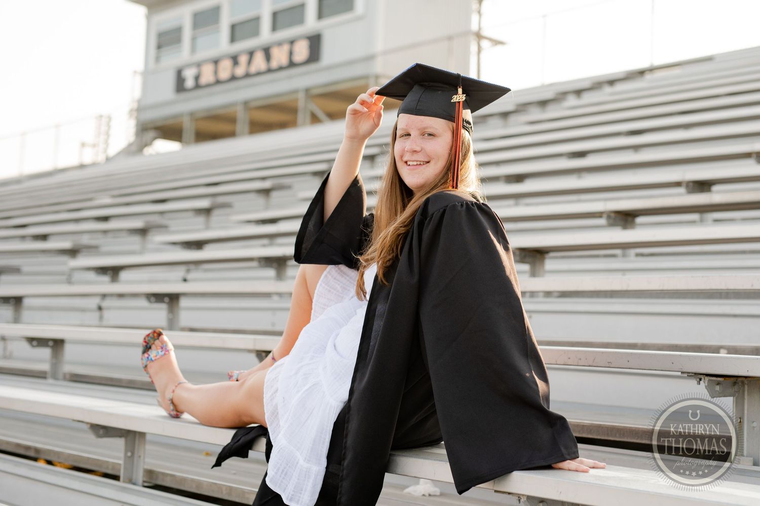 Graduate in white dress and academic regalia poses playfully on stadium bleacher steps.