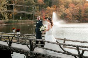 Newlyweds standing on rustic bridge with autumn foliage and fountain in background.